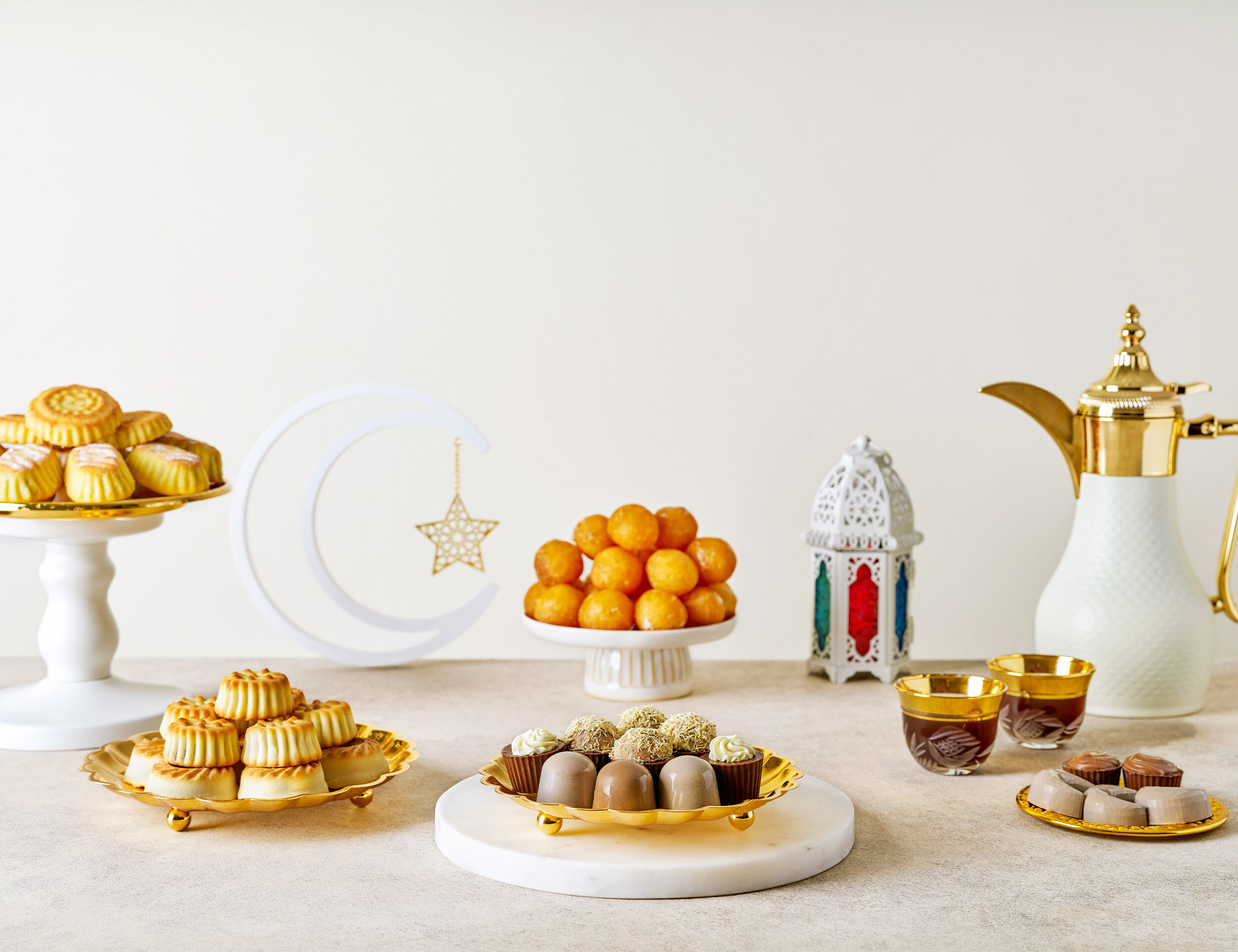 Elegant Ramadan table setting with traditional sweets, Arabic coffee, a golden dallah, and festive decorations, including a crescent moon and lantern.